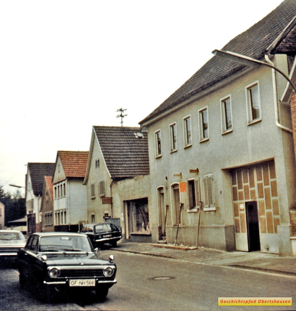 Die damalige Erzberger Straße, heute Kapellenstraße mit dem Gebäude des ehemaligen Wirtshauses Sonne, zu dieser Zeit Bäckerei Dey, Anfang der 1970er Jahre